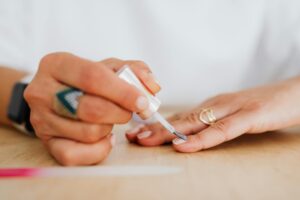 Zbliżenie na dłonie kobiety, która nakłada pędzelkiem przezroczystą odżywkę na naturalną płytkę paznokcia. Prawidłowa aplikacja preparatów stymulujących wzrost paznokci.